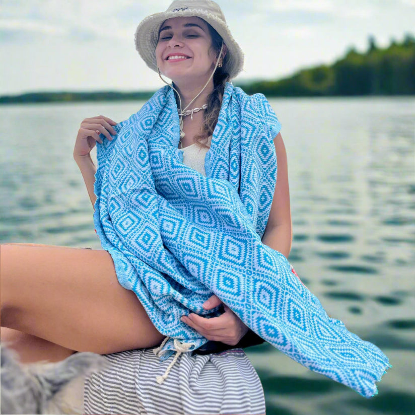 Woman sitting by a lake wearing a blue patterned towel around her shoulders.