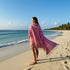 Woman walking on a beach with a pink and purple towel draped over her shoulders.